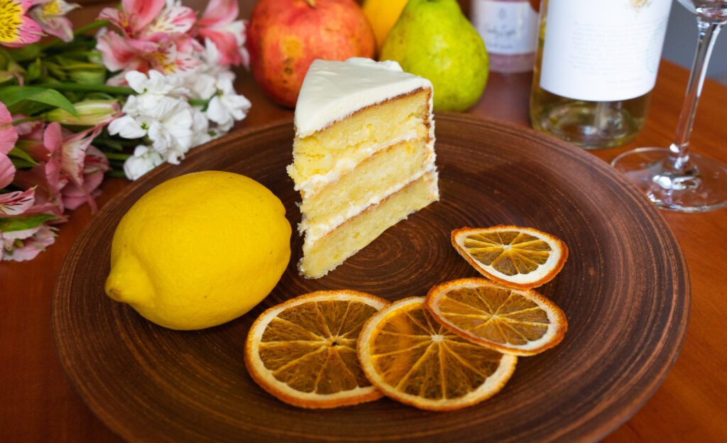 Fresh lemon cake slice on a plate with lemon and candied lemon garnish in front of a table of flowers