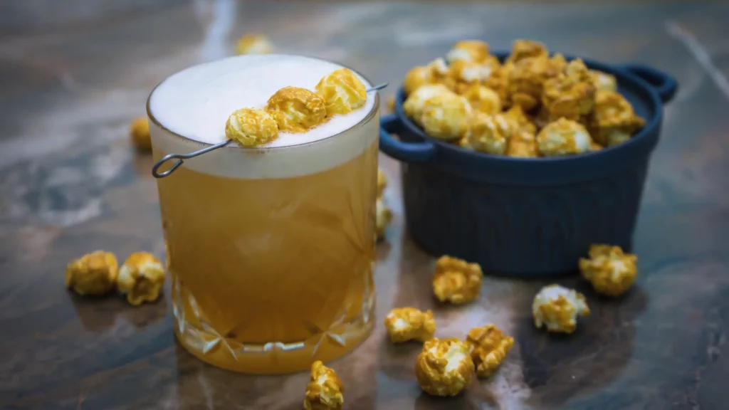 Cinema Sour cocktail next to a jar of caramel popcorn with some overflow on the marble countertop