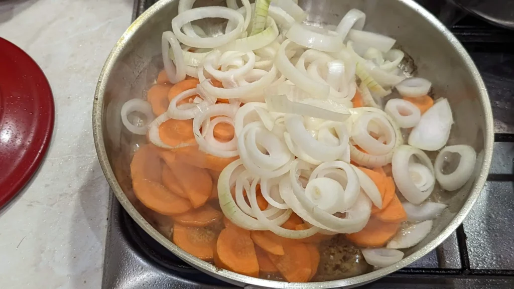 sliced onions and carrots being fried in a steel pan with duck fat on a stovetop