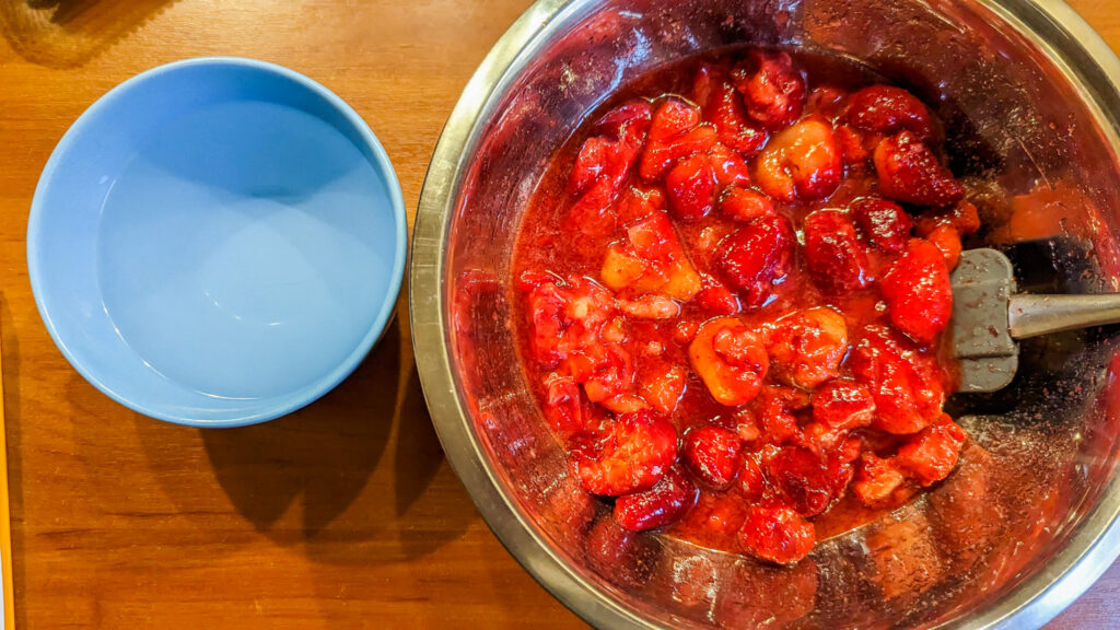 macerated strawberries in a metal bowl with a rubber spatula. Next to it on the left is a blue bowl filled with boiling water